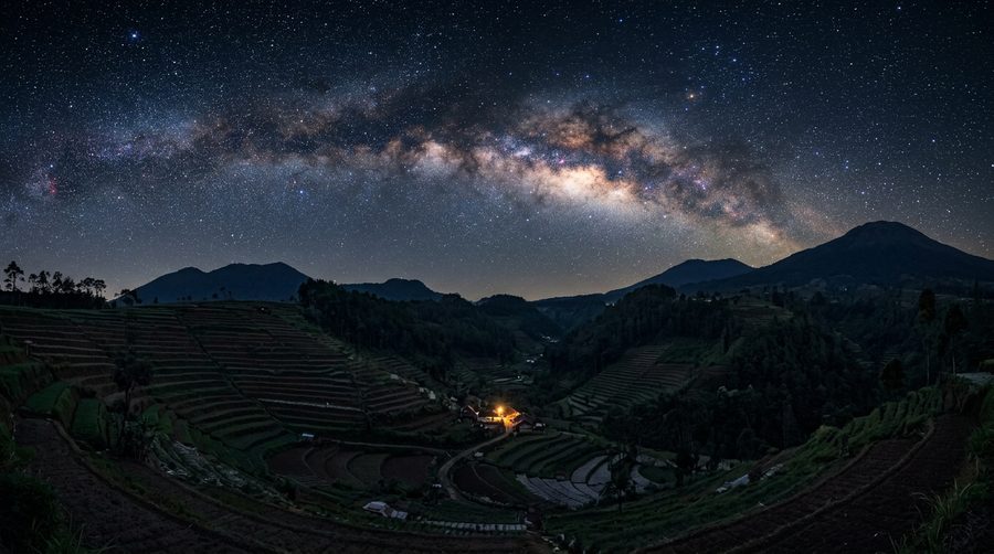 Bima Sakti membentang di langit malam gelap di atas lanskap perbukitan Indonesia dengan satu lampu hangat di kejauhan